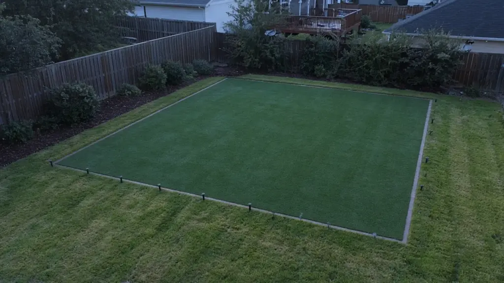 A finished artificial turf area bordered by a strip of real lawn with spray heads along the edge